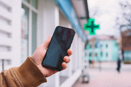 A man holds a mock-up of a smartphone on the background of a pharmacy in the city.