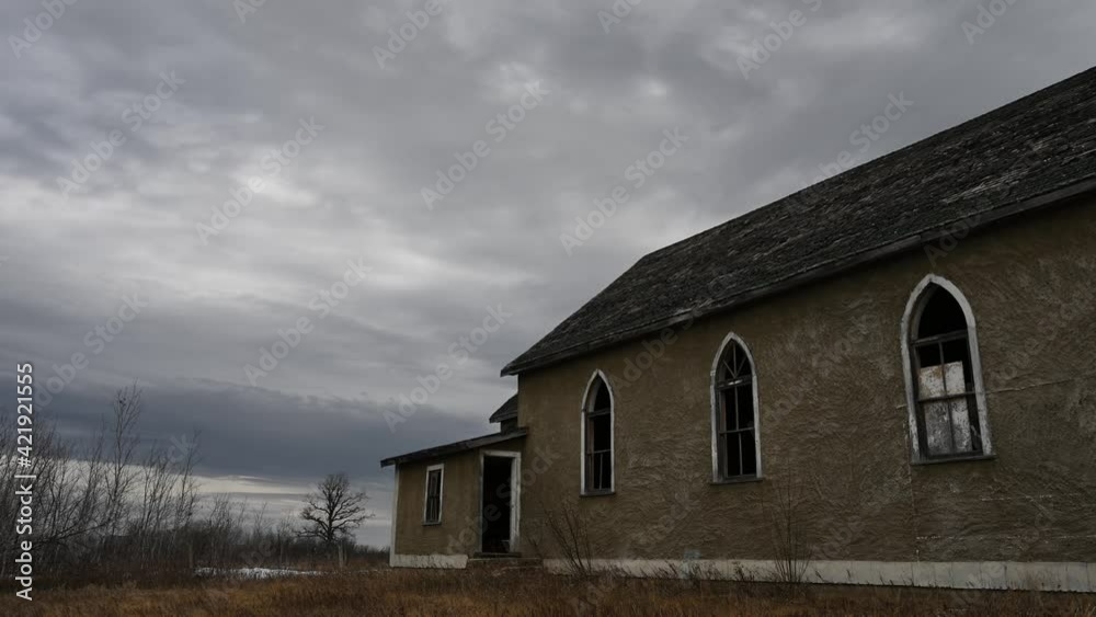 Grey clouds move over a dilapidated church with broken windows and missing shingles creating a somber scene. One leafless oak tree stands out in the distance.
Grey clouds move over a dilapidated church with broken windows and missing shingles creating a somber scene. One leafless oak tree stands out in the distance.