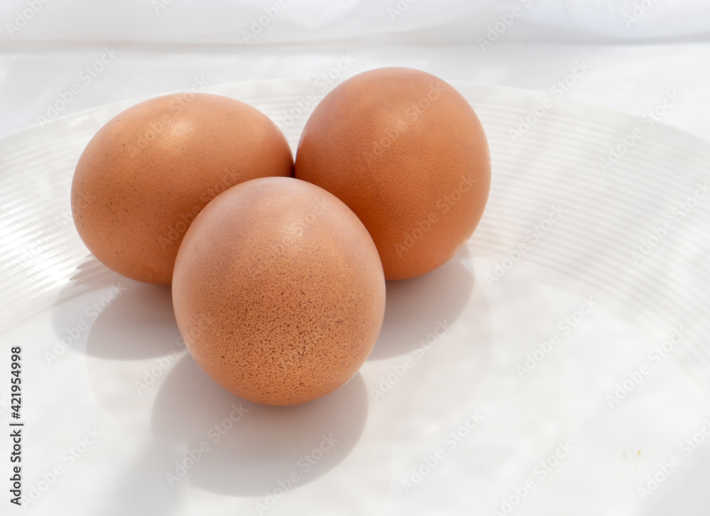 Three brown eggs placed together on a white plate and white background, with the sun coming through the window in the morning