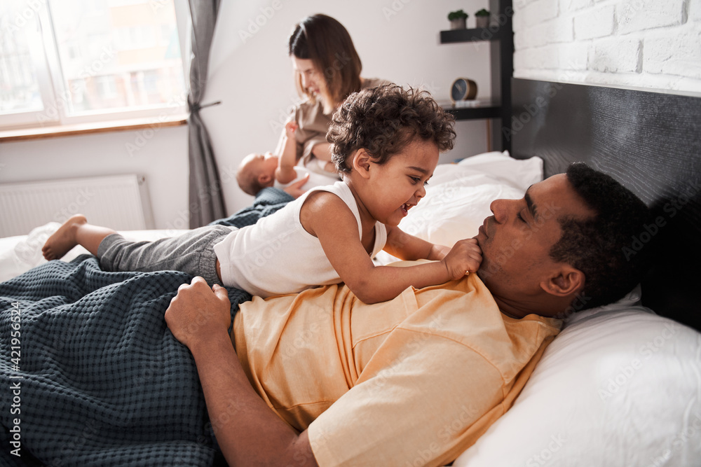 Multiracial parents and cute small kids playing while lying on bed together Multiracial parents and cute small kids playing while lying on bed together