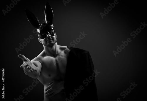 Naked topless muscular man in black rabbit mask and with jacket on shoulder stands with his arm outstretched to camera, calling us, inviting to join over dark background with copy space