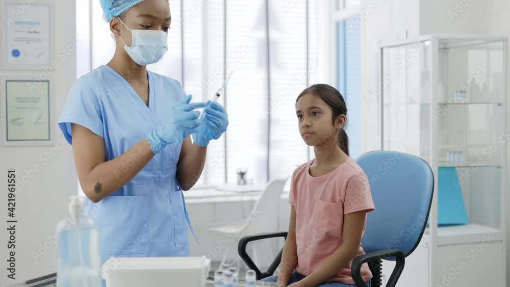 Female medical nurse in protective mask, hat and safety gloves filling syringe and prepering vaccine for young girl. Kid waiting for vaccination while sitting on chair in hospital. Female medical nurse in protective mask, hat and safety gloves filling syringe and prepering vaccine for young girl. Kid waiting for vaccination while sitting on chair in hospital.