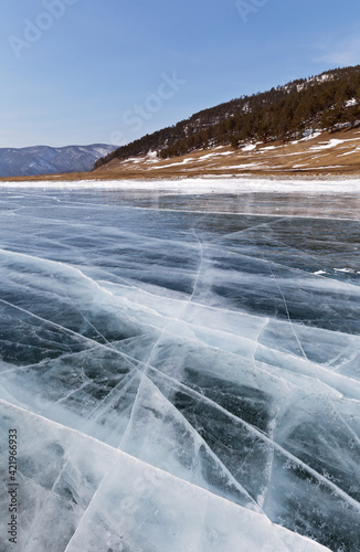 Typical winter landscape of frozen Baikal Lake with blue clear ice with cracks and coastal wooded hills on a sunny day in February. Natural background. Ice travel and outdoor holidays