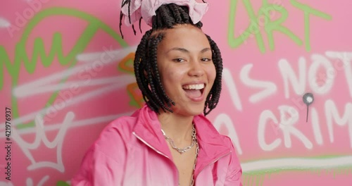 Funny woman with dreadlocks dances actively shows rap gesture shows street dance freestyle and juzz funk wears jacket has fun poses against graffiti wall. Youth curlture hip hop movement concept photo