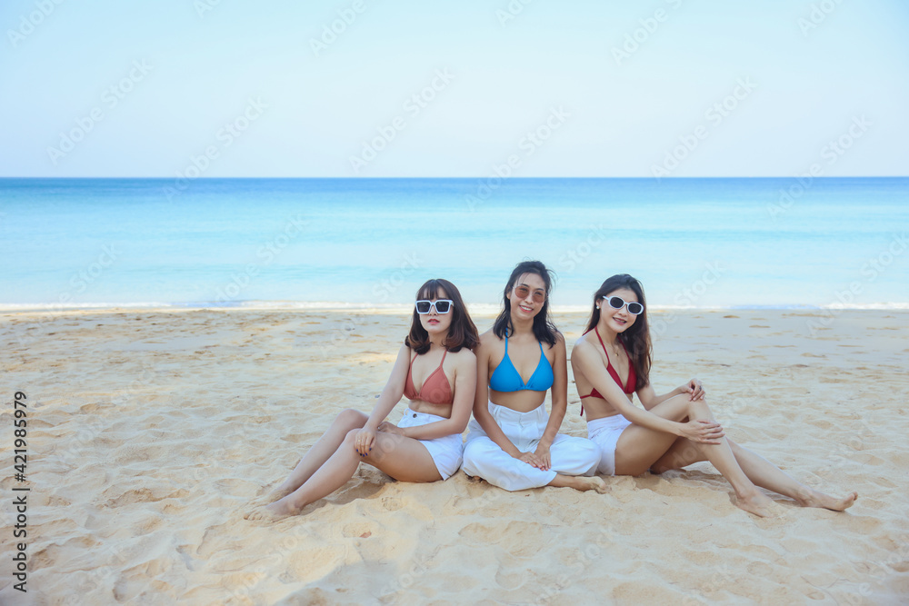Portrait of three beautiful young asian women friends, wearing bikini and stylish sunglasses sitting on the seashore looking at camera smile. Blue sea and sky in the background Portrait of three beautiful young asian women friends, wearing bikini and stylish sunglasses sitting on the seashore looking at camera smile. Blue sea and sky in the background