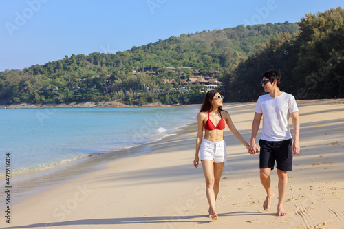 Portrait of beautiful young women and boyfriend asian, wearing rea bikini and stylish white sunglasses walking on the seashore looking at camera smile. Blue sea and sky in the background