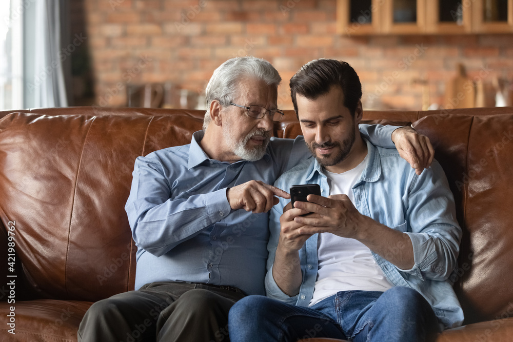 Mature Caucasian father and adult son relax on sofa at home using modern smartphone gadget. Old 60s dad and grownup man kid rest on couch in living room browse internet on cellphone device.