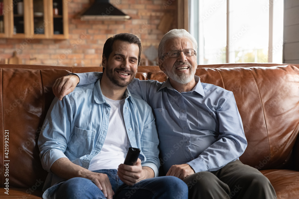 Happy elderly Caucasian grey-haired father and adult 20s son sit relax on couch at home watch TV together. Smiling mature dad and 30s grownup man child rest on sofa in living room enjoy television.