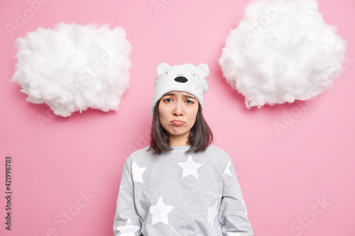 Sad brunette Asian woman looks dissatisfied at camera has sulking face expressionn being offended by someone feels lonely to spend time at home wears slumber suit bear hat isolated on pink wall photo