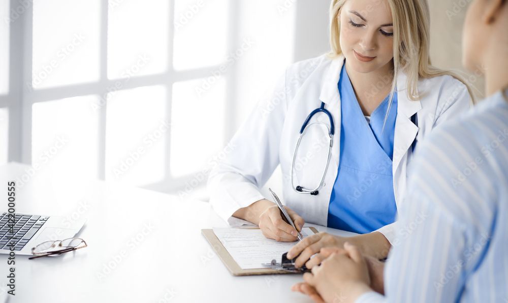 Woman-doctor and patient sitting and talking in hospital or clinic. Blonde therapist is cheerfully smiling. Medicine concept Woman-doctor and patient sitting and talking in hospital or clinic. Blonde therapist is cheerfully smiling. Medicine concept