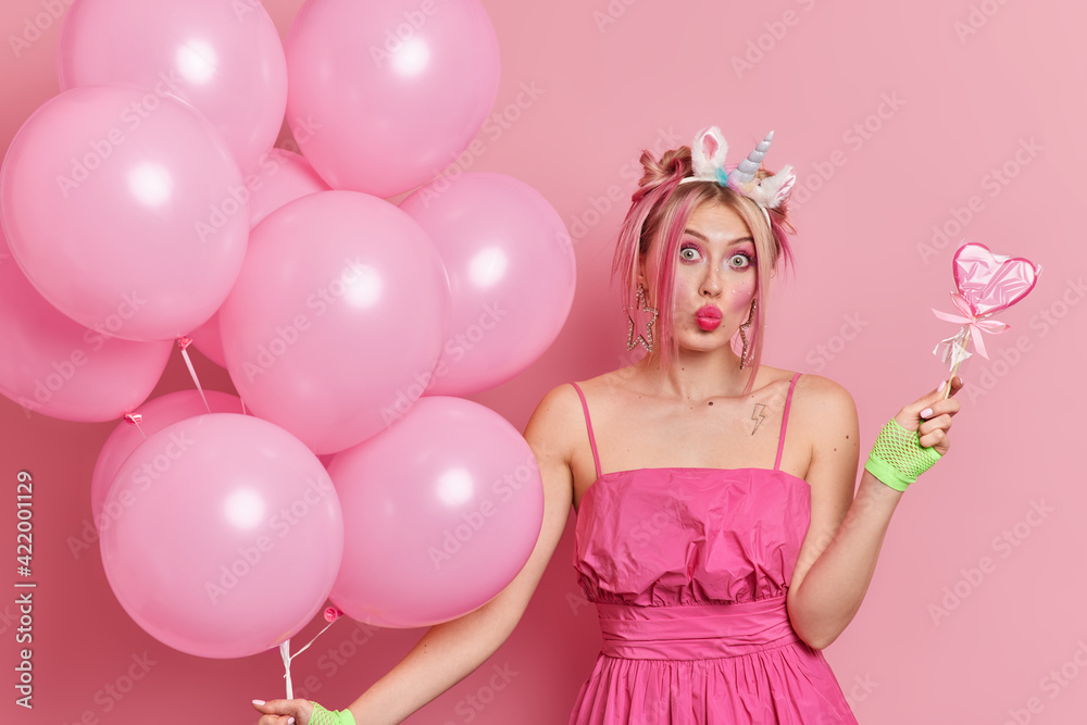 Stylish surprised European woman folds lips has fun on birthday party holds delicious candy and bunch of inflated balloons has great mood isolated over pink background. Festive occasion concept Stylish surprised European woman folds lips has fun on birthday party holds delicious candy and bunch of inflated balloons has great mood isolated over pink background. Festive occasion concept