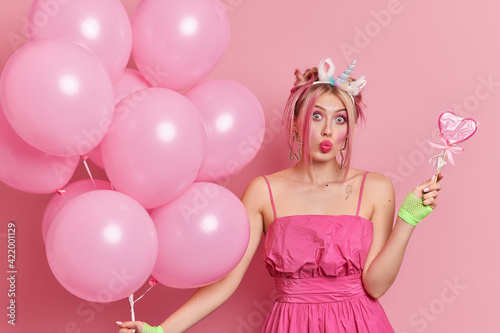 Stylish surprised European woman folds lips has fun on birthday party holds delicious candy and bunch of inflated balloons has great mood isolated over pink background. Festive occasion concept