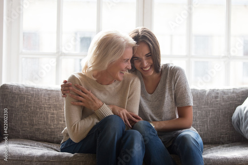Smiling adult daughter and mature 60s mother sit relax on sofa at home have fun laugh enjoy family time together. Happy millennial grownup woman child cuddle hug with old mom on couch, show love care. photo
