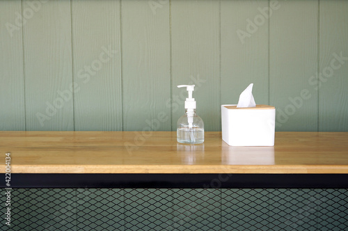 Tissue paper in a white box and hand alcohol gel bottle are on a wooden table. Showing hygiene and germs protection in house, restaurant, working space during pandemic. Grey panels in background.