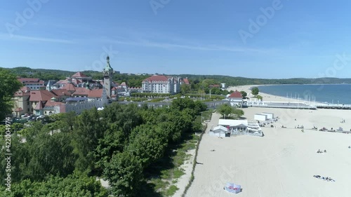 Sandy beach leading straight down to the blue sea. In the middle of the frame there is a boulevard with lots of green trees. Sopot in Poland visible on a video from a drone.