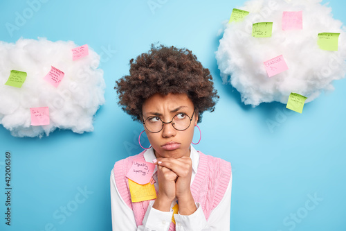 Serious thoughtful female business worker makes shorts notes on stickers to memorize keeps hands under chin wears eround spectacles neat clothes isolated over blue background prepares for workshop photo
