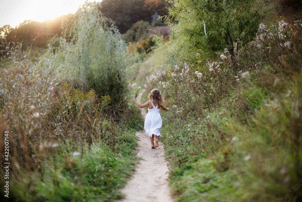 A young blonde girl in a white dress runs along the path in the park barefoot, happy free childhood, closeness to nature, natural and careful upbringing