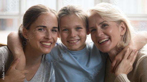Headshot portrait of smiling small Caucasian girl child hug happy older grandmother and young mother. Close up of overjoyed three generations of women embrace cuddle show family unity and support.