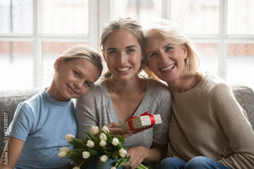 Portrait of smiling three generations of Caucasian women relax at home celebrate anniversary together. Happy little girl child daughter and grandmother congratulate young mother with gift and flowers.