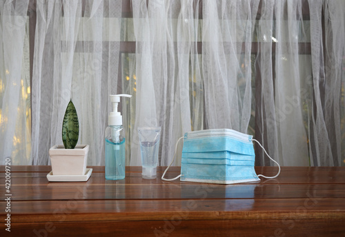 A blue alcohol pump, a hand gel sanitizer and a face mask are on dark wood table.  All items are important for people during Covid 19 pandemic. White curtain in blurred background.