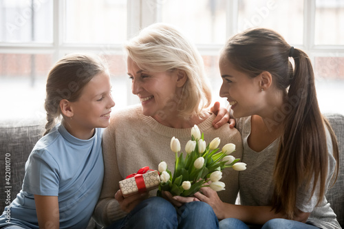 Cute small Caucasian girl child and young mother greeting excited senior grandmother with birthday anniversary present gift flowers. Three generations of women celebrate special occasion together.