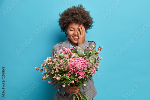 People holiday event concept. Positive Afro American woman holds beautiful bouquet of flowers received on birthday covers face with hand dressed in grey jacket isolated over blue background.