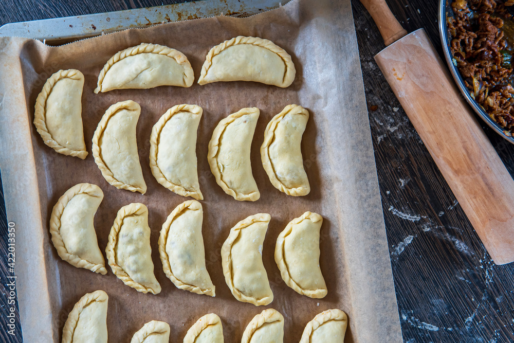 Fototapeta premium Uncooked empanadas on a baking tray. Traditional Latin American baked beef pastry ready for the oven. Gluten free savory appetizer with meat filling. Handmade typical dish in Spain or Argentina. Top.
