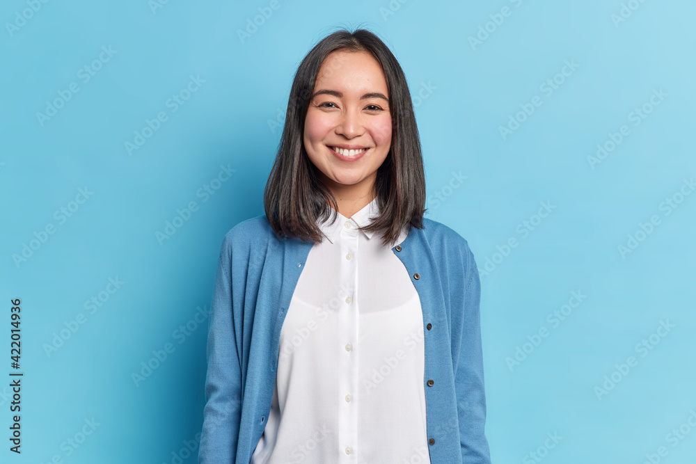 Horizontal shot of pretty Asian woman with dark hair smiles pleasantly looks directly at camera has toothy smile wears white shirt and jumper isolated over blue background. Emotions concept. Horizontal shot of pretty Asian woman with dark hair smiles pleasantly looks directly at camera has toothy smile wears white shirt and jumper isolated over blue background. Emotions concept.