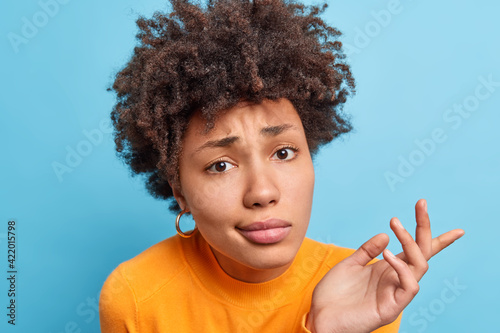 Close up shot of curly haired confused woman shrugs shoulders with hesitation has dissatisfied face expression wears orange jumper isolated over blue background being doubtful. Hard decision