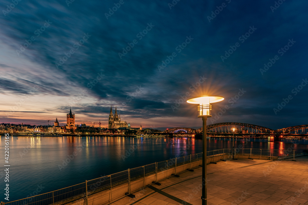 View of Cologne Cathedral with Hohenzollern Bridge at nightfall and a lamppost in the foreground, Germany.