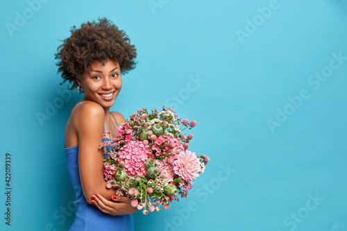 Sideways shot of young African American woman wears dress holds bouquet of flowers gets it as present on 8 March poses against blue background with copy space for your promotion. Spring concept