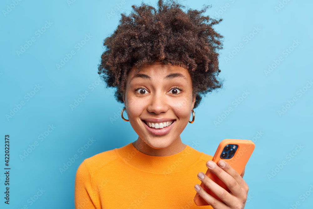 Portrait of good looking smiling young woman with curly hair uses mobile phone for chatting online downloads new app looks gladfully at camera wears orange jumper isolated over blue background