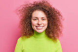 Portrait of good looking cheerful European woman with curly bushy hair smiles broadly dressed in green turtleneck isolated over pink background. Carefree smiling European female enjoys free time