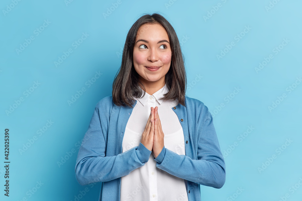 Hopeful beautiful Asian woman presses palms together prays for something makes supplicating namaste pray gesture dressed in neat clothes isolated over blue background being thankful for help Hopeful beautiful Asian woman presses palms together prays for something makes supplicating namaste pray gesture dressed in neat clothes isolated over blue background being thankful for help