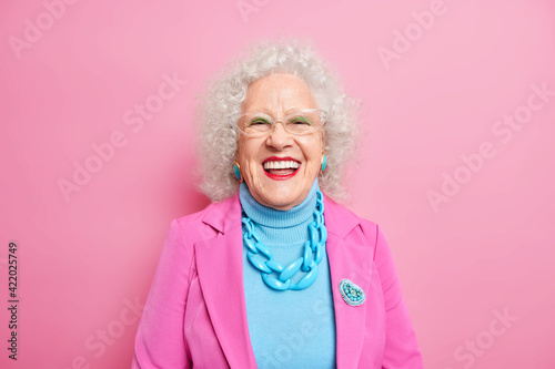 Portrait of aged beautiful woman with curly grey hair bright makeup smiles happily expresses positive emotions dressed in fashionable outfit isolated over pink background. Positive grandmother photo