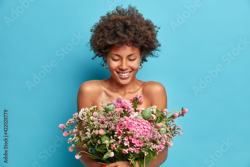 Photo of good looking cheerful curly haired Afro American woman poses with bare shoulders gets present from beloved person enjoys spring time has happy mood isolated over blue studio background.