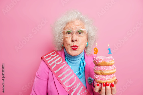 Portrait of beautiful female pensioner keeps lips rounded wants to kiss you and says thankful for congratulations being well dressed holds pile of delicious doughnuts isolated over pink background