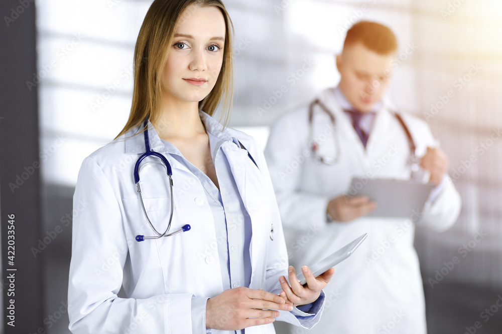 Professional woman doctor with a stethoscope is using a computer tablet, while she is standing together with her colleague in a sunny clinic. Young doctors at work in a hospital. Medicine concept Professional woman doctor with a stethoscope is using a computer tablet, while she is standing together with her colleague in a sunny clinic. Young doctors at work in a hospital. Medicine concept