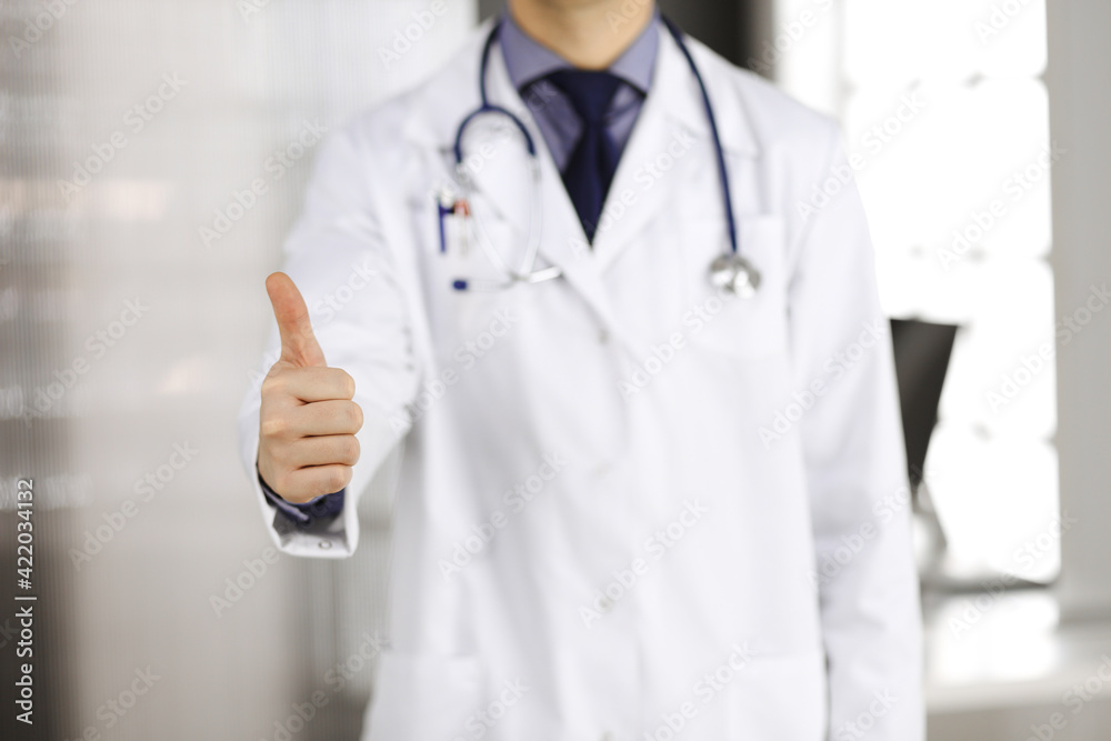 Unknown male doctor standing with thumbs up sign in clinic near his working place, closeup. Perfect medical service in hospital. Medicine and healthcare concept Unknown male doctor standing with thumbs up sign in clinic near his working place, closeup. Perfect medical service in hospital. Medicine and healthcare concept