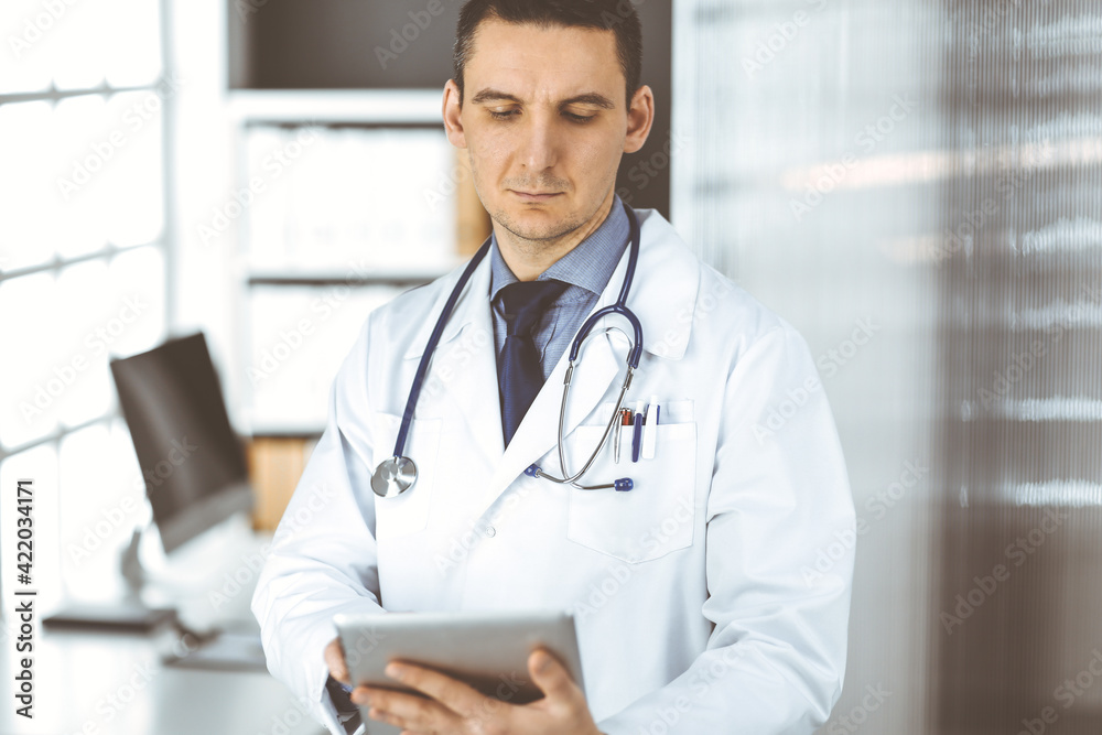 Friendly male doctor using tablet computer in clinic near his working place. Perfect medical service in hospital. Medicine and healthcare concept Friendly male doctor using tablet computer in clinic near his working place. Perfect medical service in hospital. Medicine and healthcare concept