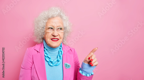 Mature beautiful woman with wrinkled skin wears stylish clothes jewelry indicates at upper right corner give wide advice shows copy space against pink backgroud. People age advertisement concept
