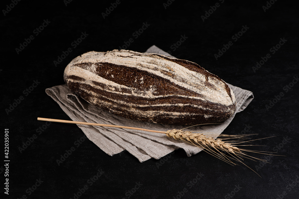 Artisan sourdough rye bread on dark background. Whole loaf of bread on textile napkin and rye spikelet.
