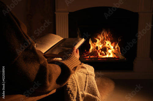 Woman reading book near fireplace indoors, closeup. Cozy atmosphere