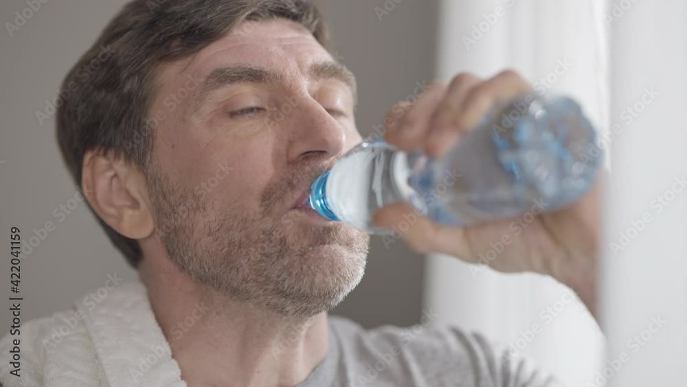 Headshot portrait of perspiring man wiping forehead with hand, drinking refreshing water from bottle and looking at camera smiling. Close-up of handsome middle aged Caucasian sportsman. Headshot portrait of perspiring man wiping forehead with hand, drinking refreshing water from bottle and looking at camera smiling. Close-up of handsome middle aged Caucasian sportsman.