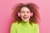 Close up shot of overjoyed curly haired woman laughs out smiles broadly has curly bushy hair listens music via wireless stereo headphones dressed in green turtleneck isolated on pink background