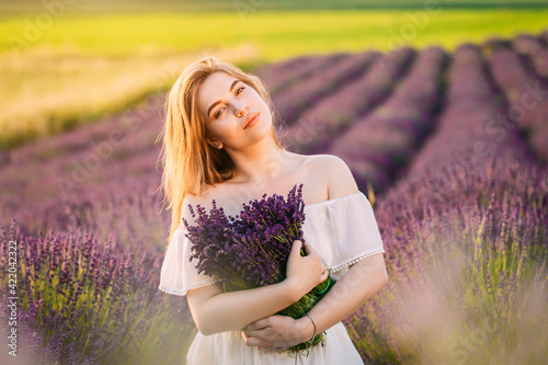 Young and happy blond woman and white dress enjoying spring in a lavender field at sunset holding a bouquet of flowers in her hands