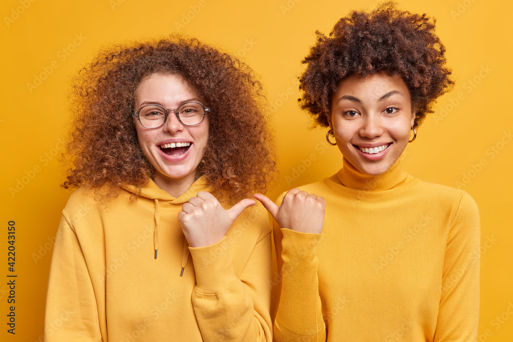 Φωτογραφία Happy joyful mixed race curly haired women point at each other with cheerful expression say its she stand closely to each other dressed casually isolated over yellow background