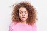 Headshot of stunned woman with curly bushy hair stares with disbelief at camera reacts on amazing news wears casual clothes isolated over white background. Human reaction and emotions concept