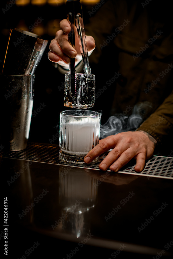 bartender neatly holds large piece of ice with tongs over steaming old-fashioned glass