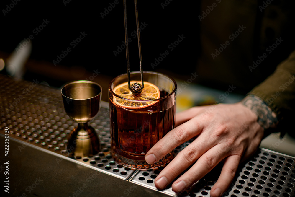 man decorating by small pine cone an old-fashioned glass of cocktail with orange slice man decorating by small pine cone an old-fashioned glass of cocktail with orange slice